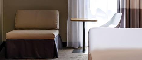 A hotel room with a bed, a sofa, a small table, and a view of the cityscape through the window.