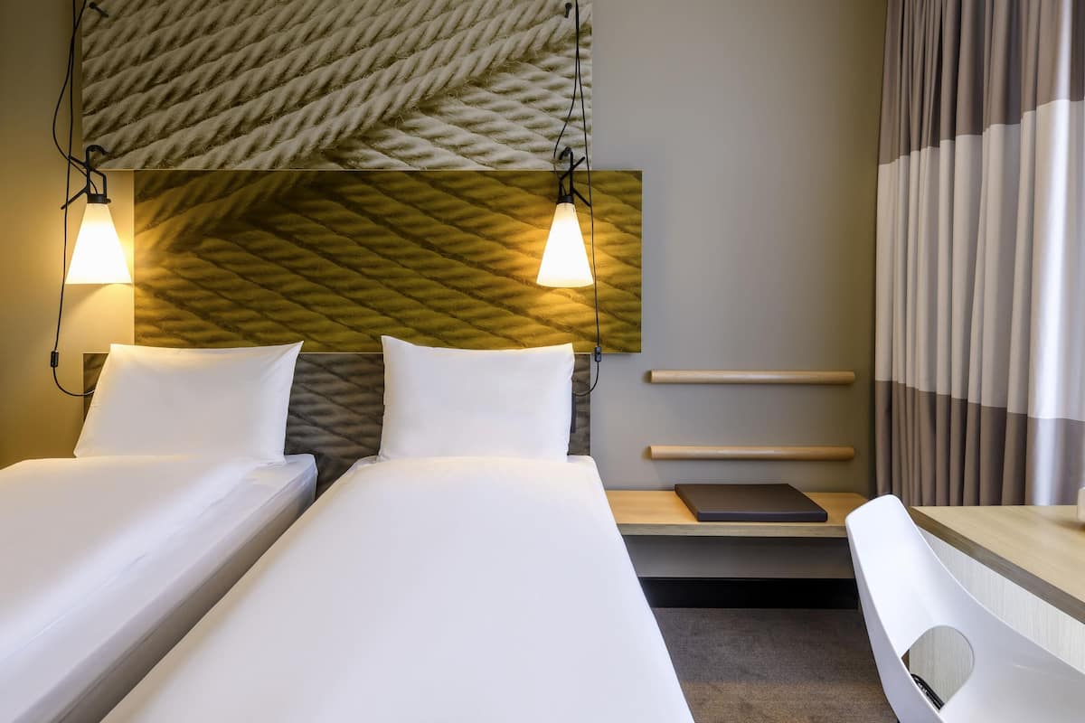 Habitació d'hotel amb dos llits, un capçal de llit amb dibuixos en relleu, làmpades de nit, una taula petita i una cadira.