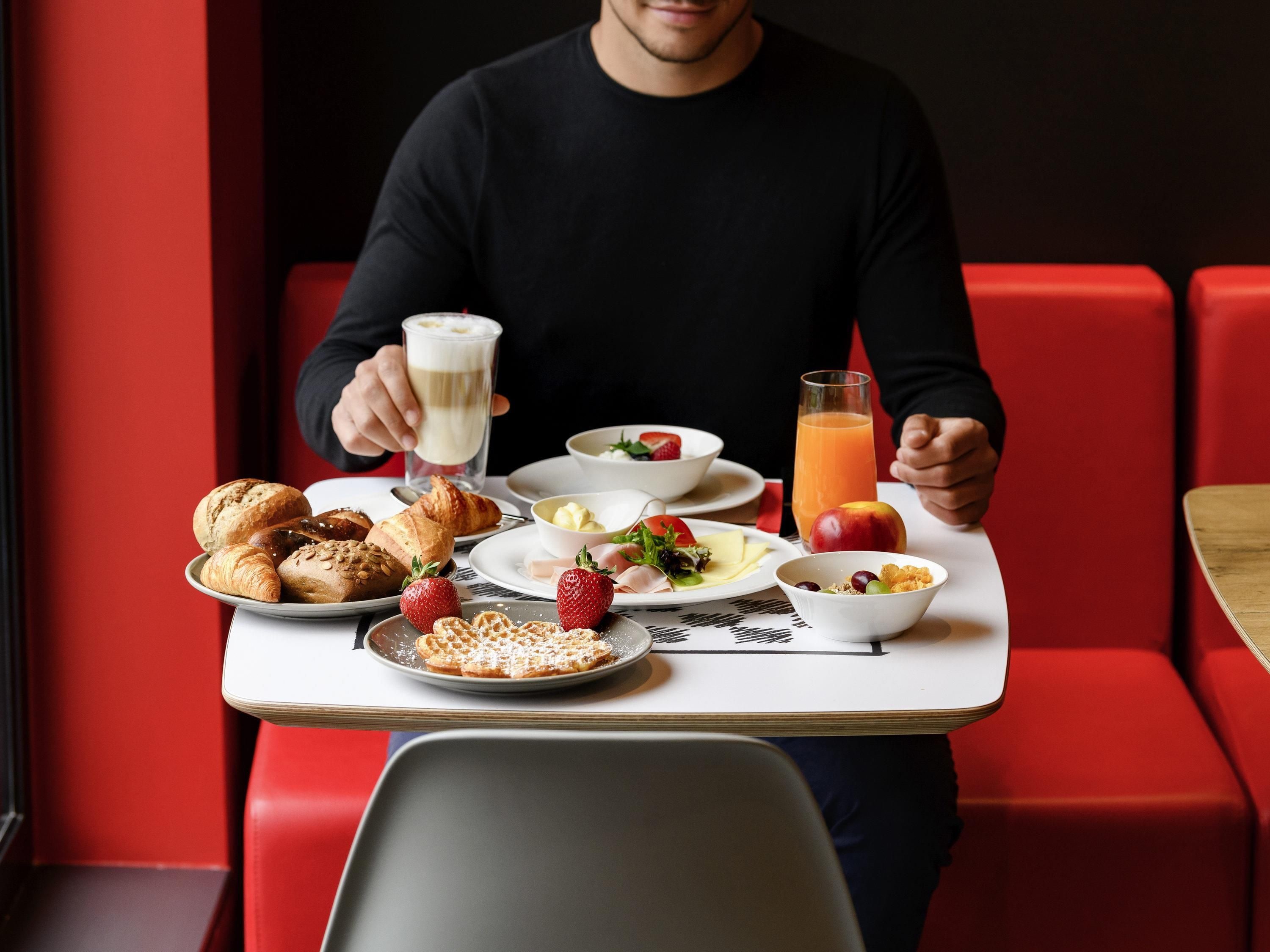 Una persona sentada a una mesa con alimentos para el desayuno, como bollería, fruta y bebidas.