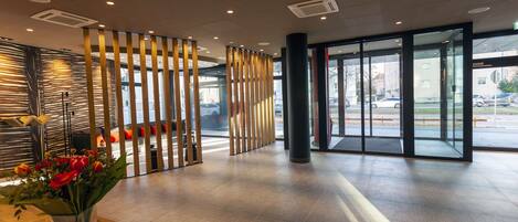 A modern lobby with a reception desk, a glass partition with vertical blinds, and a view of the street outside.