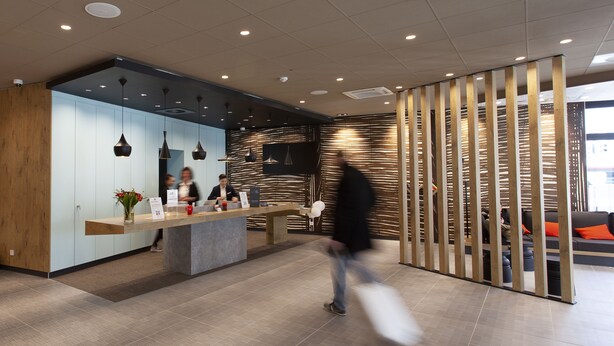Een moderne receptiezone met een houten receptiebalie, een zithoek met oranje kussens, en een scheidingswand met verticale lamellen.