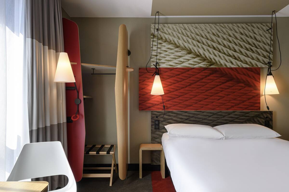 Habitación de hotel con cama, lámparas de noche, pared roja, cabecero de madera, una mesita, una silla y una ventana con cortinas.