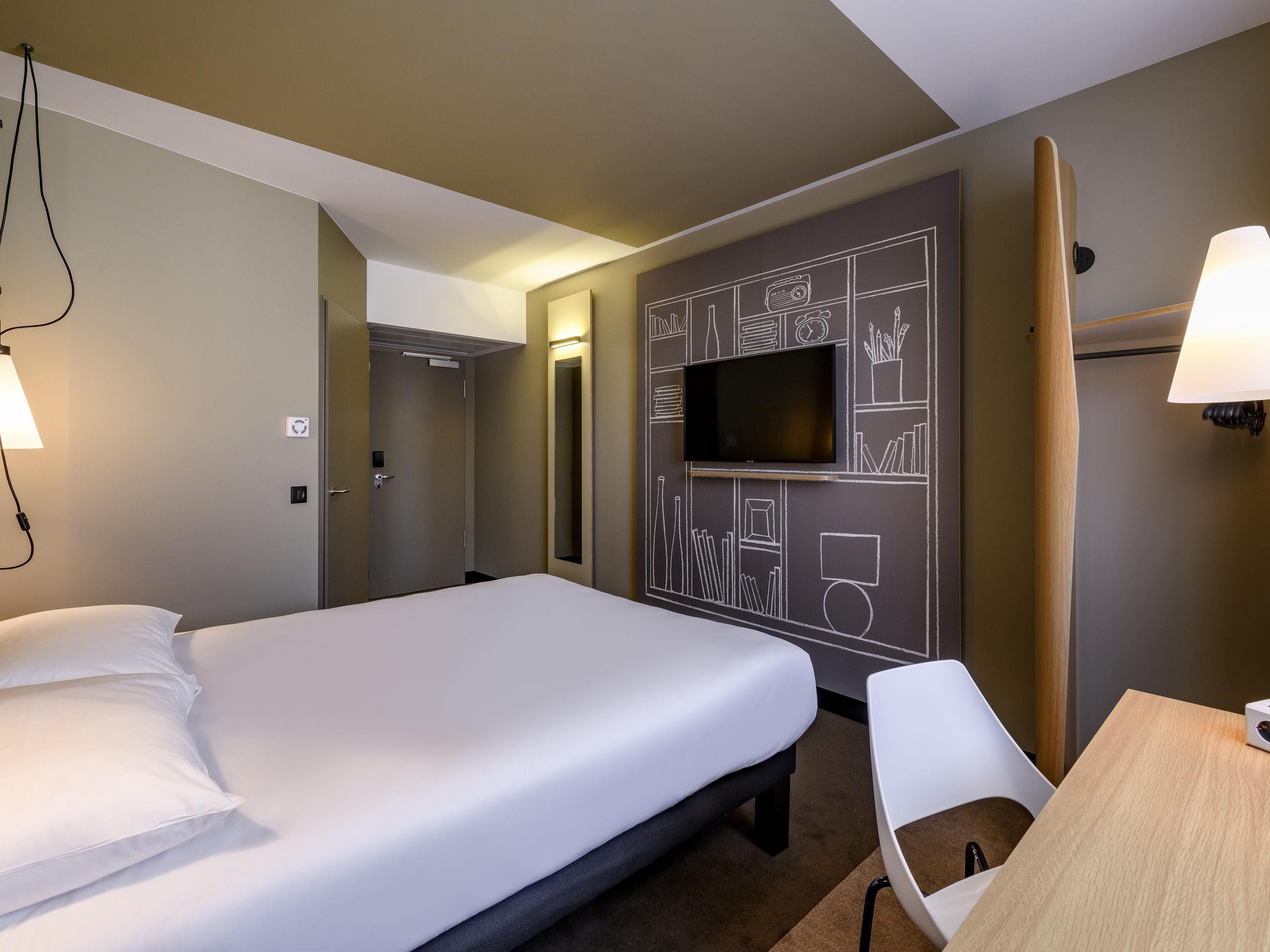 Una habitación de hotel con una cama, un televisor montado en la pared, un escritorio con una silla y una puerta que da a otra habitación.