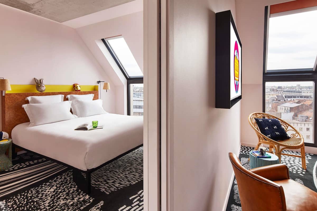 A hotel room with a bed, a small table, a chair, a window with a city view, and a skylight.