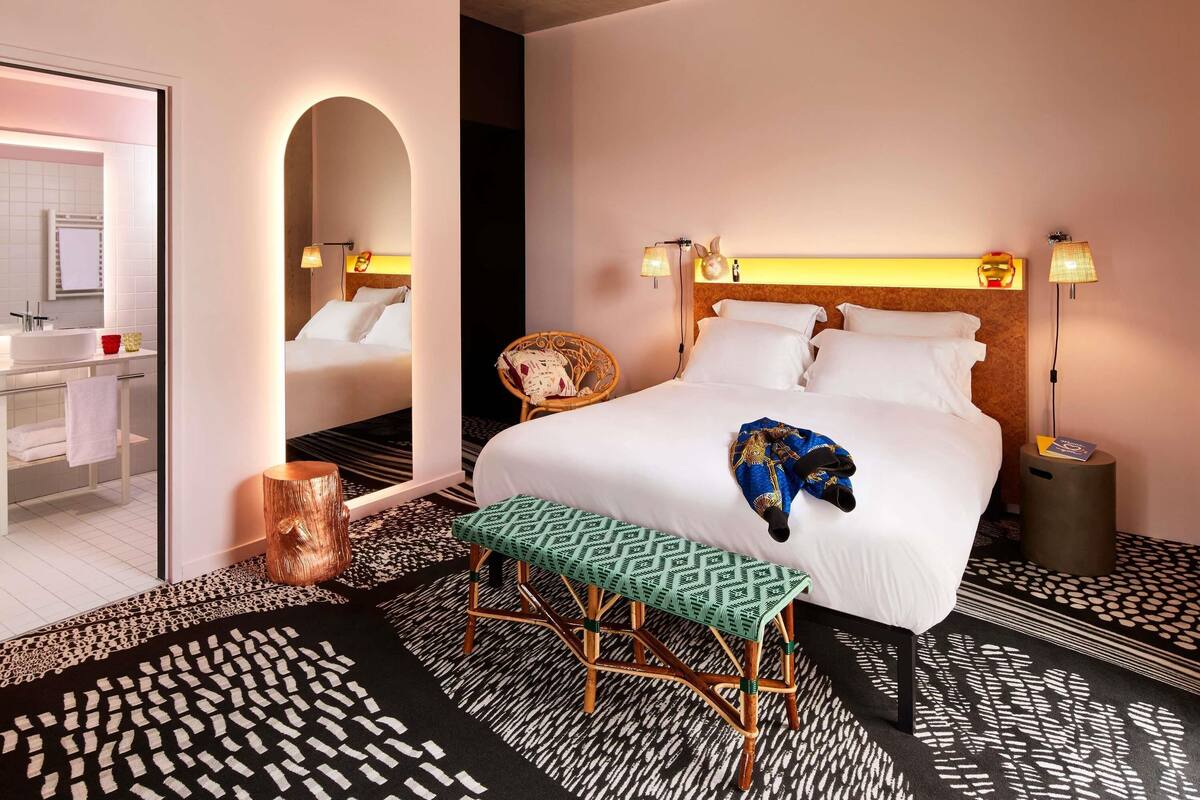 A hotel room with a bed, bedside lamps, a bench, a chair, a mirror, and a bathroom visible through an open door.