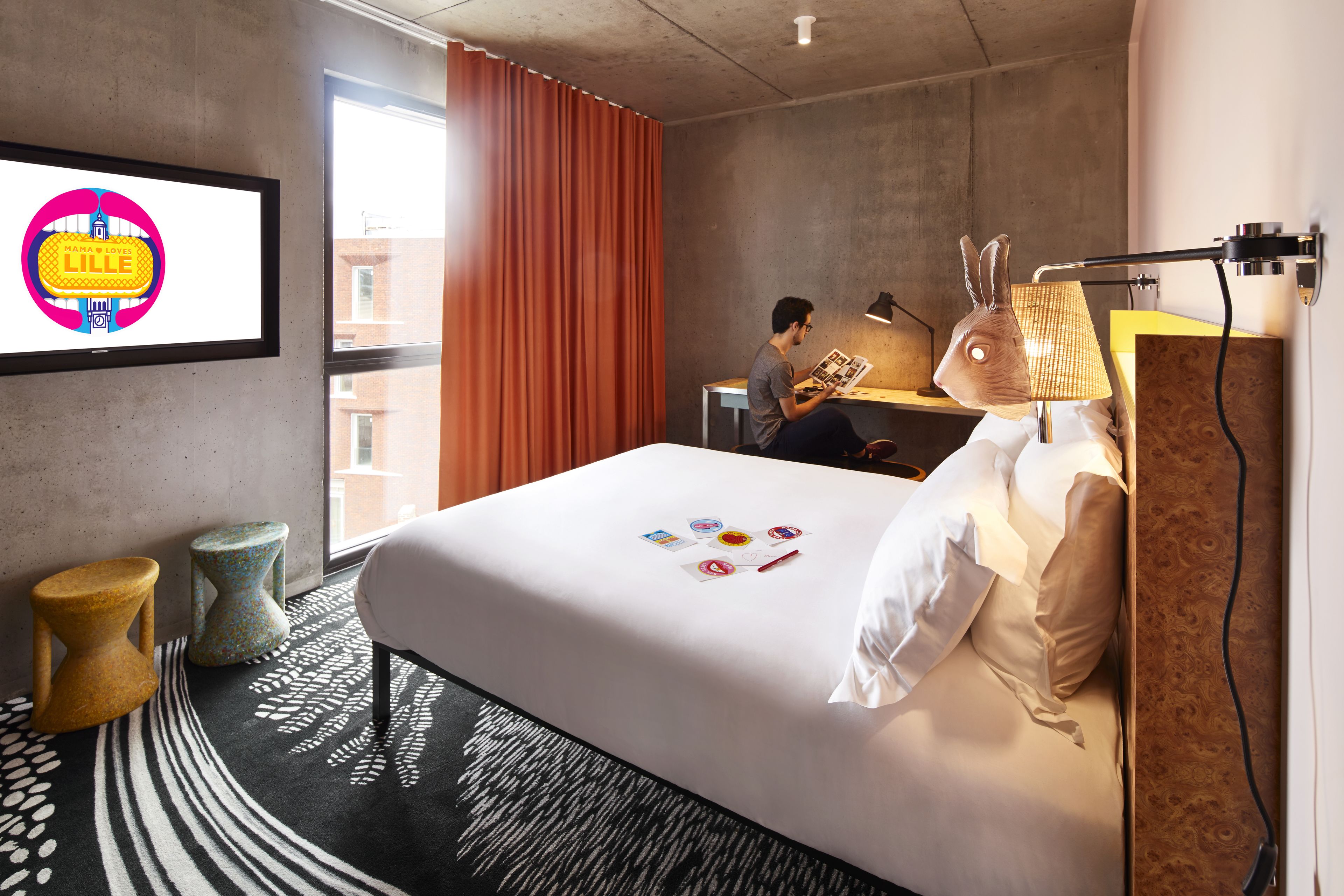 A hotel room with a bed, a desk, a laptop, a lamp with a rabbit head, and a wall-mounted TV.