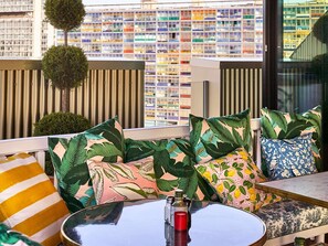 A modern outdoor seating area with patterned cushions, a round table, and a view of a colorful building in the background. - Mama Shelter Lille (Lille)