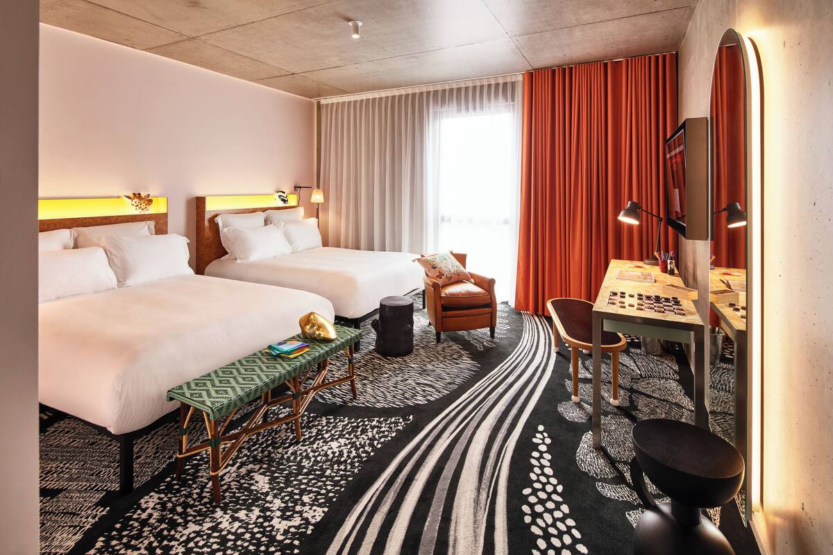 A hotel room with two beds, a desk with a chair, a small table, and a large mirror.