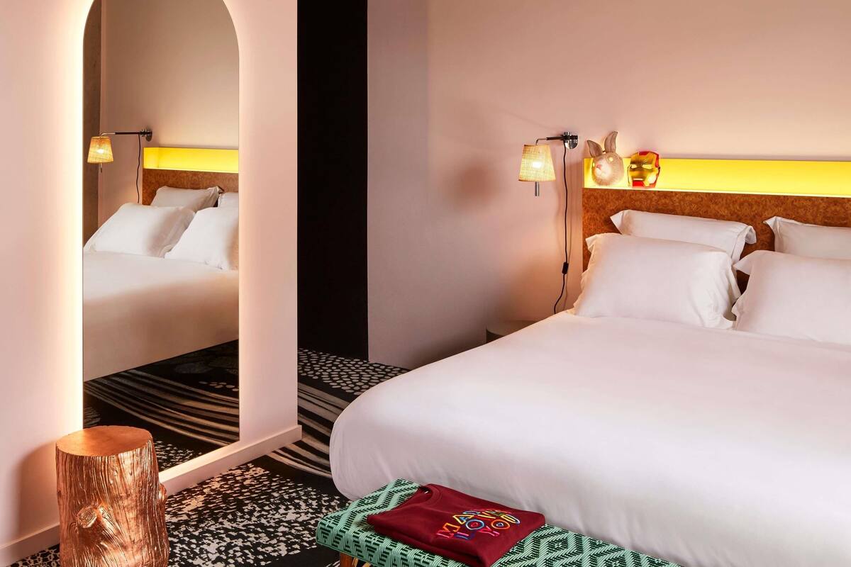 A hotel room with two beds, a mirror, a wooden stool, and a patterned carpet.