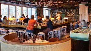 Uma área de bar com banquetas, mesas e uma mesa de pebolim.