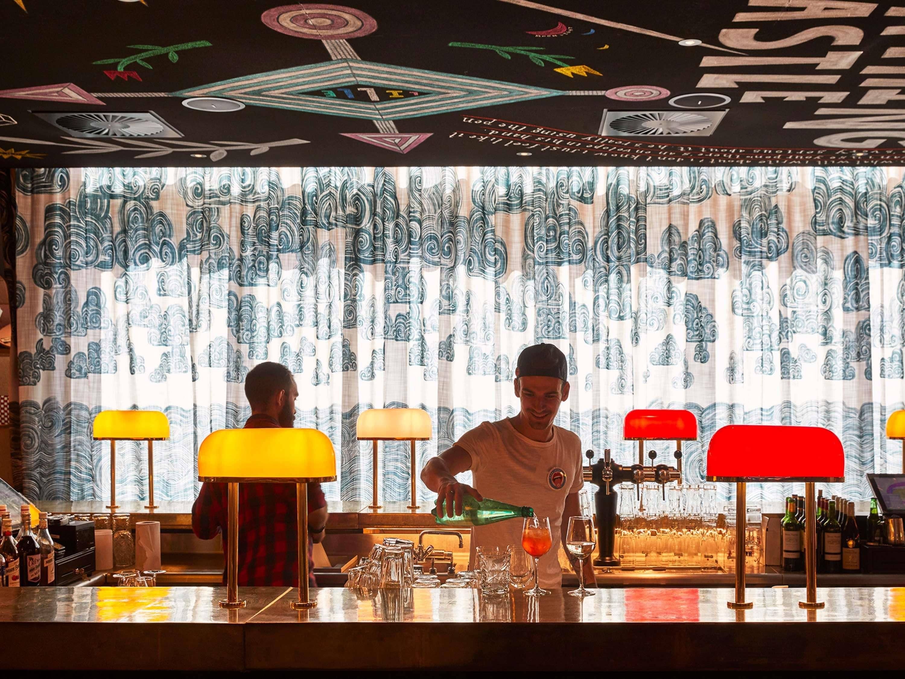 Un espace bar agrémenté d’un rideau décoratif à motifs, de tabourets de bar et d’un barman préparant des boissons.