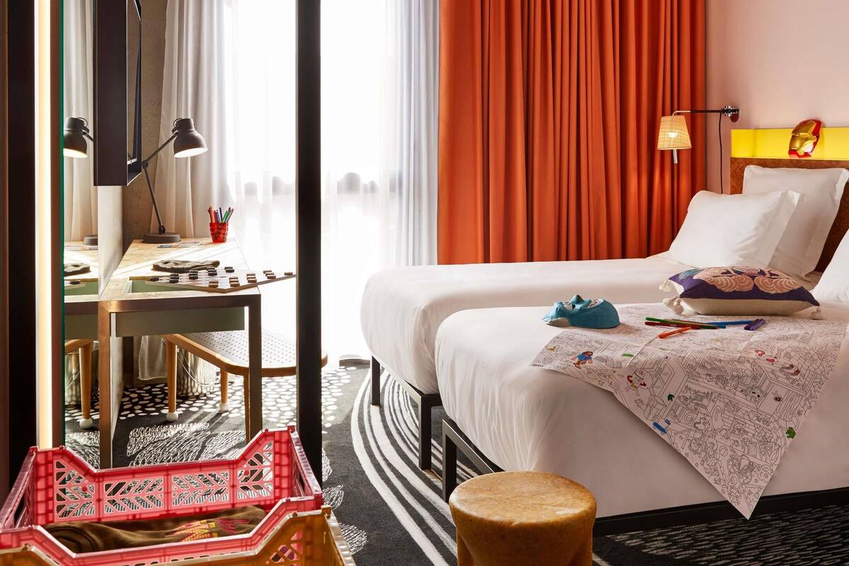 A hotel room with a bed, a desk with a lamp, a chair, a small stool, and a window with curtains.