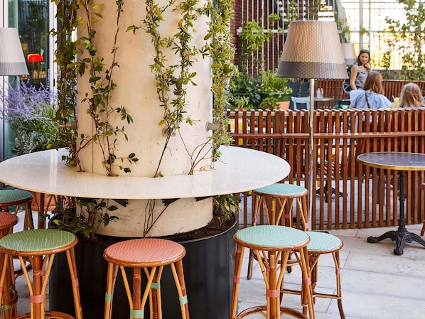 A round table with a central column, surrounded by wicker chairs with green cushions, in an outdoor seating area with a wooden fence and potted plants.