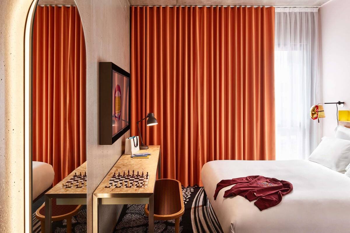 A hotel room with a bed, a desk with chess, a mirror, curtains, and a lamp.