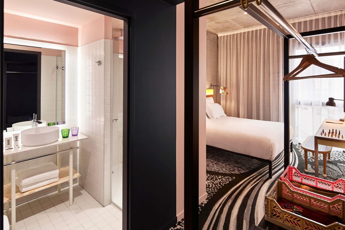 Une chambre d’hôtel avec salle de bain, un lit, un bureau avec un jeu d’échecs et une vue sur la ville.