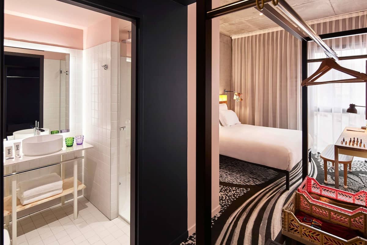 A hotel room with a bathroom, a bed, a desk with a chessboard, and a view of the city.