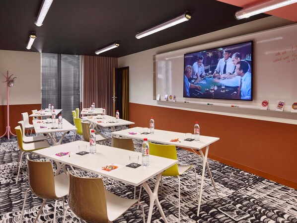 A conference room with tables, chairs, a projector screen, and a television displaying a group discussion. - Mama Shelter Lille (Lille)