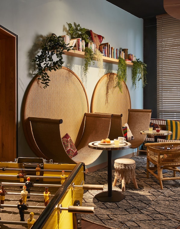 A cozy lounge area with rattan furniture, a small table with drinks, and a bookshelf with books and plants. - Mama Shelter Paris West (Paris)