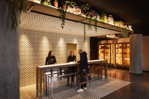 An indoor area with a patterned wall, a reception desk, and shelves with plants and books. - Mama Shelter Paris West (Paris)