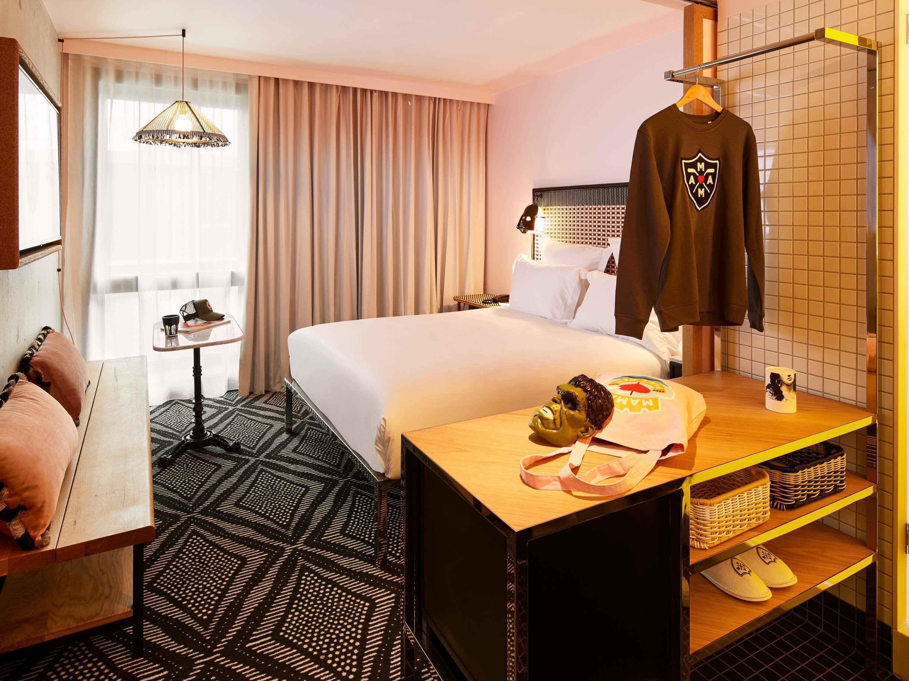 A hotel room with a bed, a desk with a mug, a hanging shirt, and a mirror.