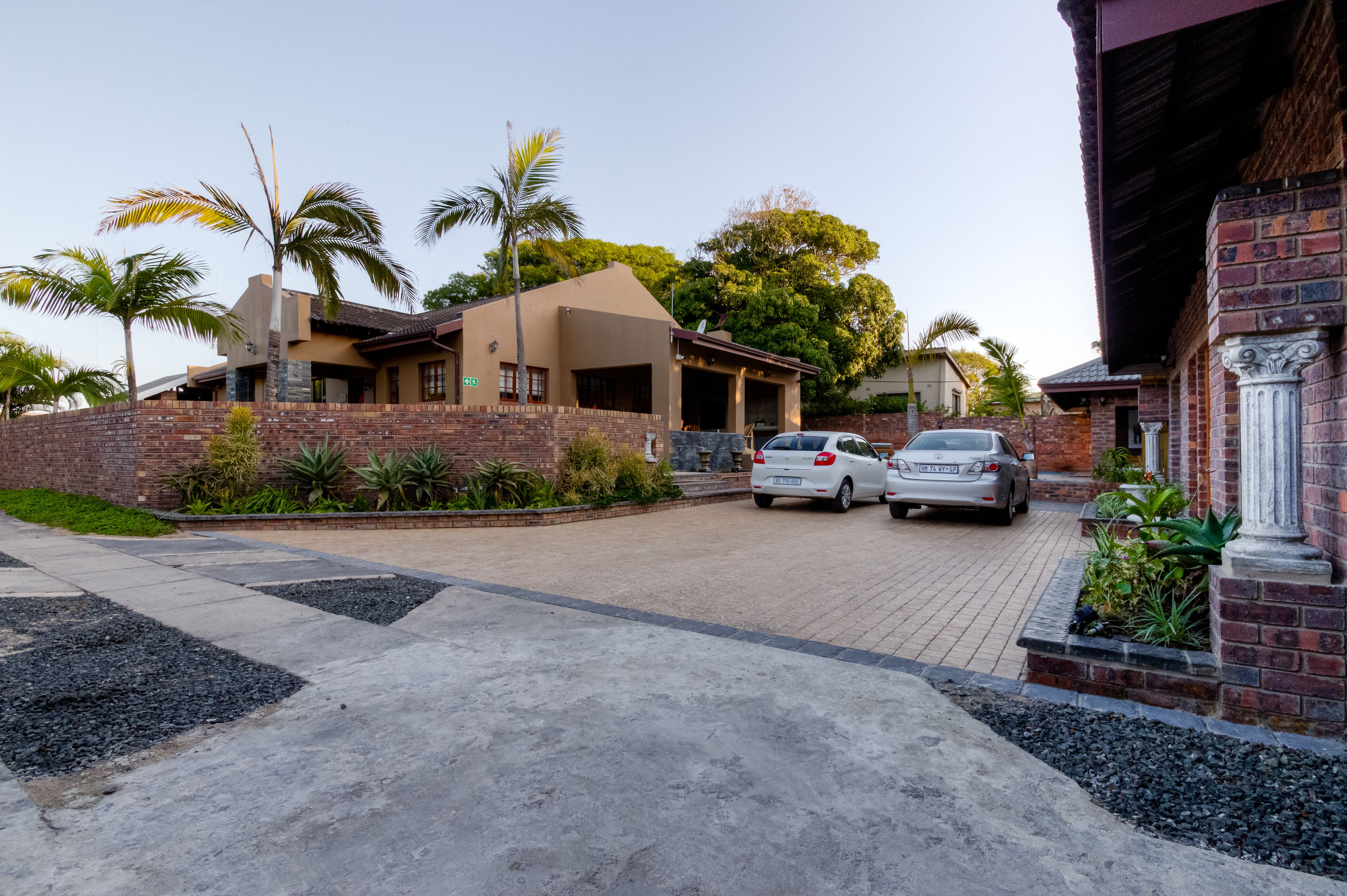 A residential area with two cars parked in a driveway, a brick wall, and a house with a sloped roof.