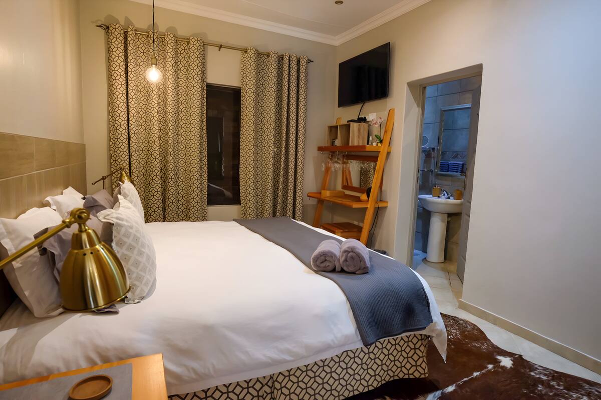 Quarto de hotel com cama, luminária de mesa, banheiro com pia e televisão fixada na parede.