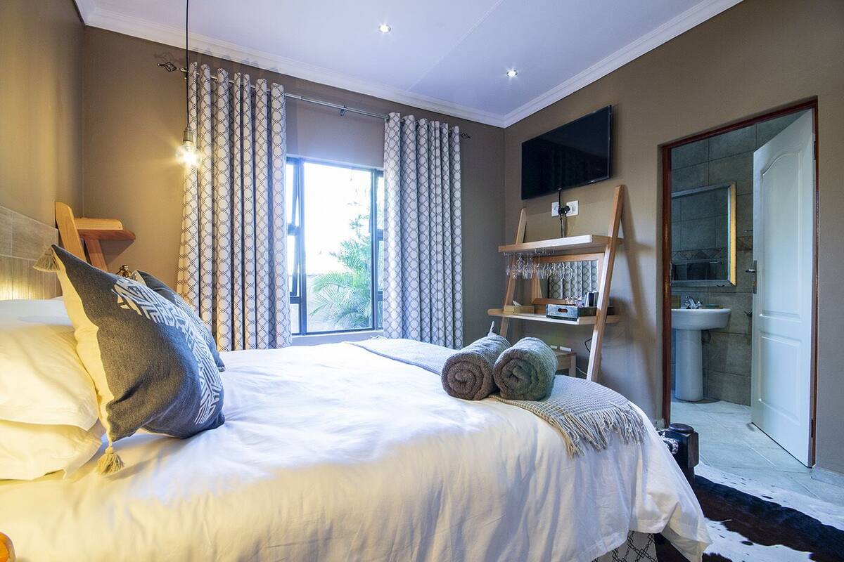 Quarto com cama grande, televisão na parede, prateleira de madeira com copos e vista para o exterior pela janela.