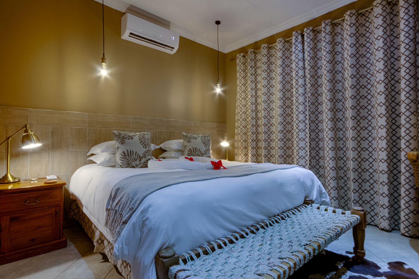 A hotel room with a bed, bedside lamps, an air conditioner, patterned curtains, and a bench.