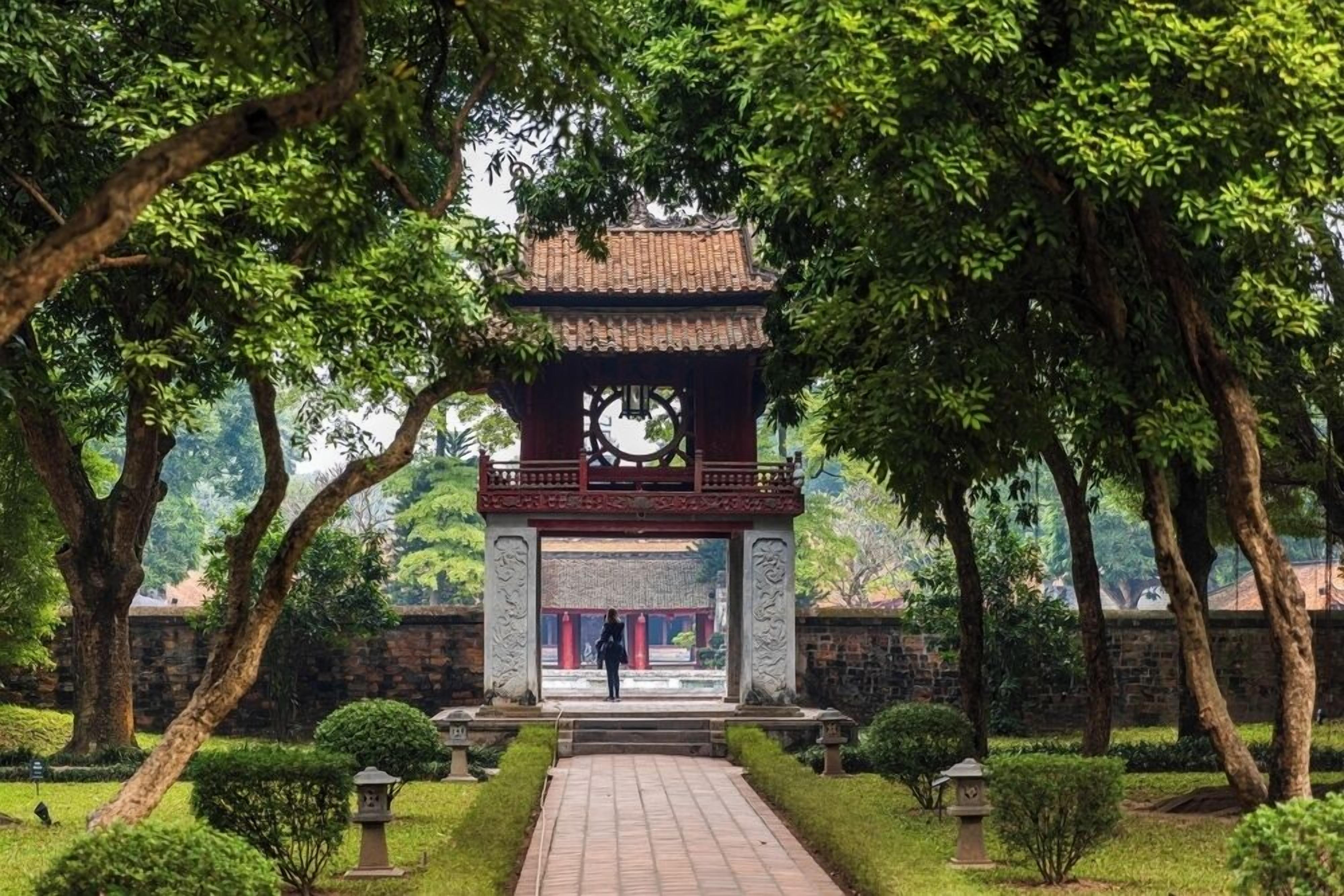 A traditional pavilion with a tiled roof, surrounded by lush greenery and a stone wall.