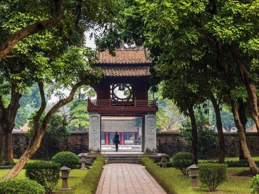 A traditional pavilion with a tiled roof, surrounded by lush greenery and a stone wall.