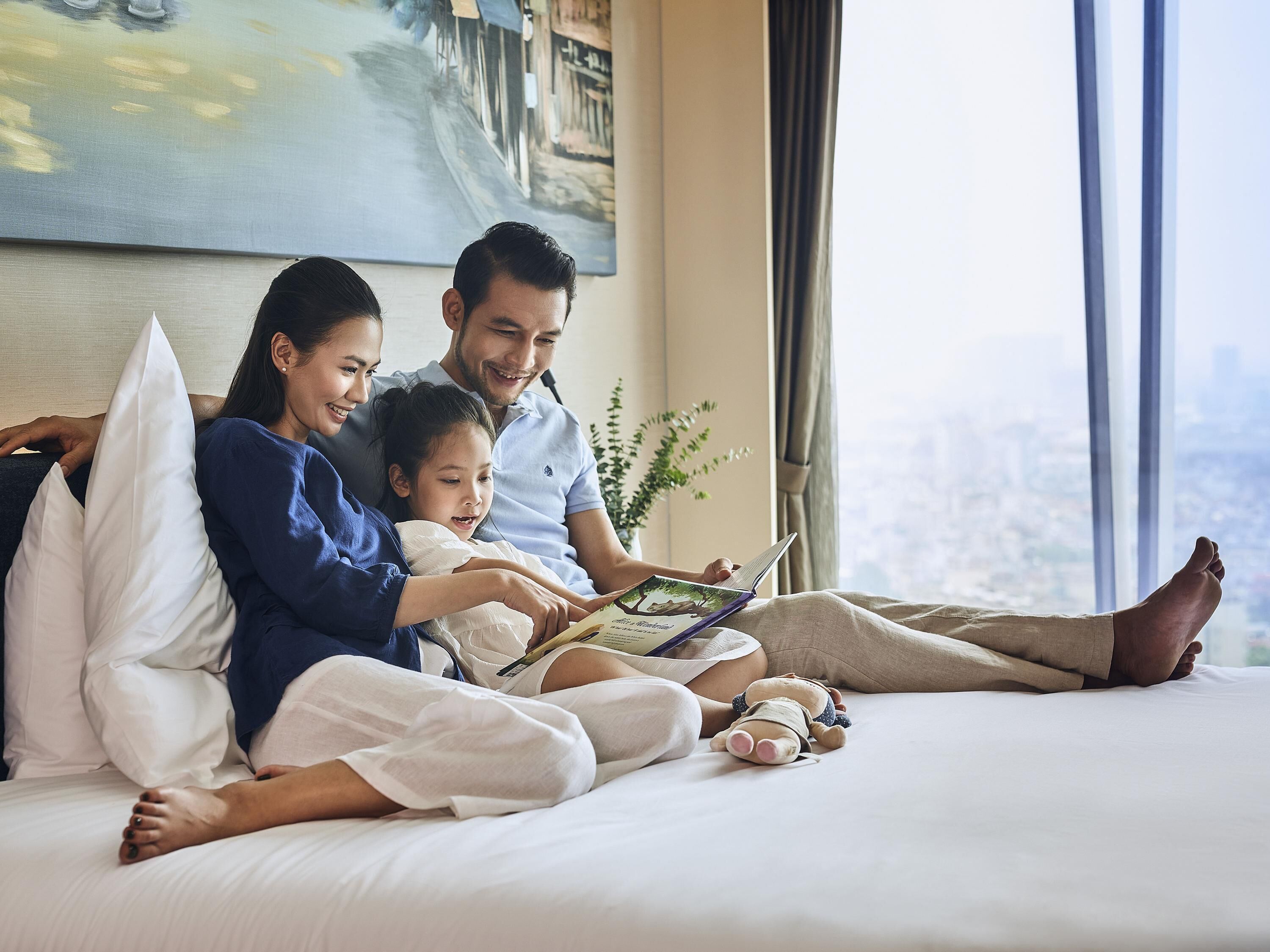 Una familia de tres en una cama con vista a la ciudad, un cuadro en la pared y una planta junto a la ventana.