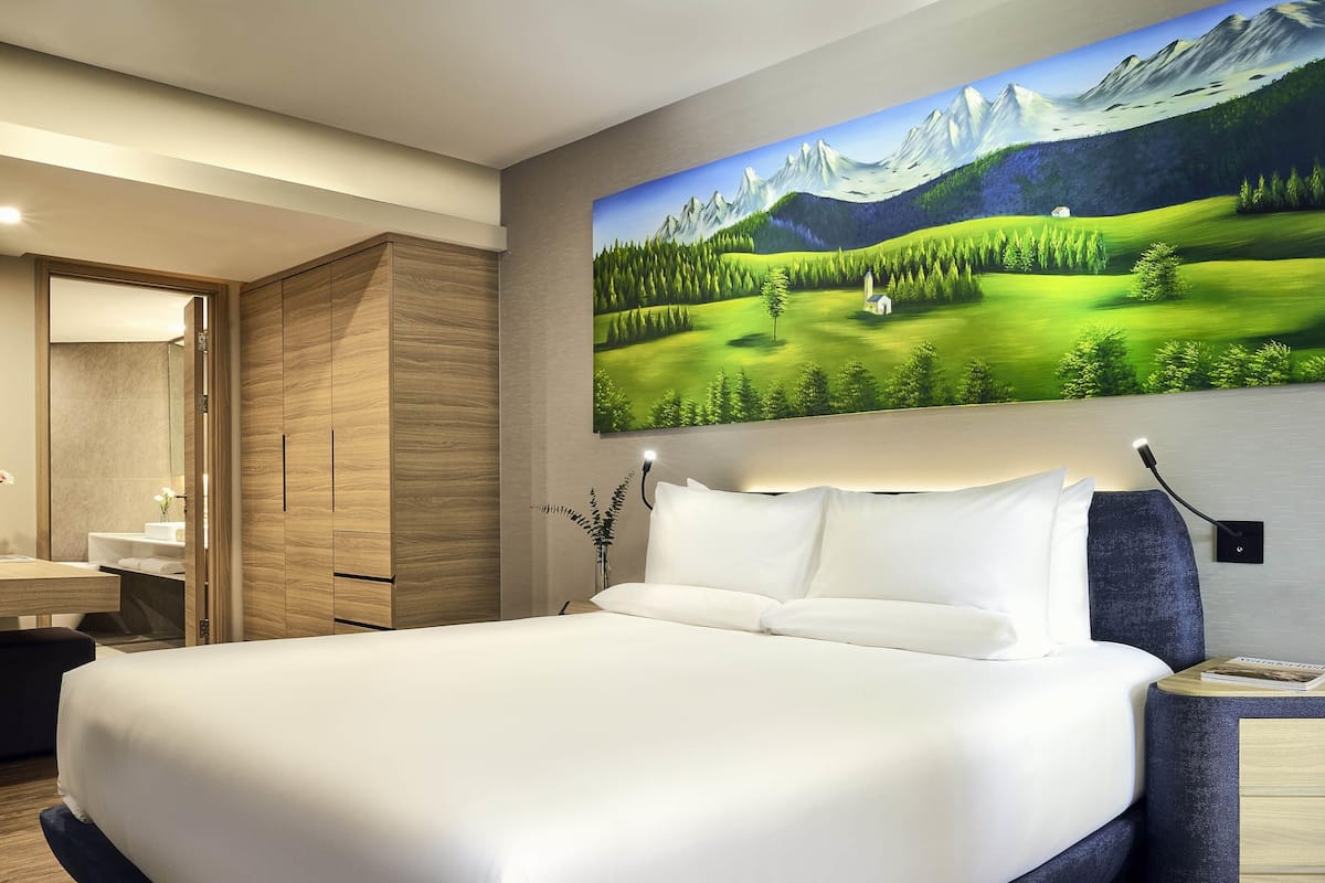 A modern hotel room with a large bed, a wooden wardrobe, a bathroom with a mirror, and a painting of a mountainous landscape.