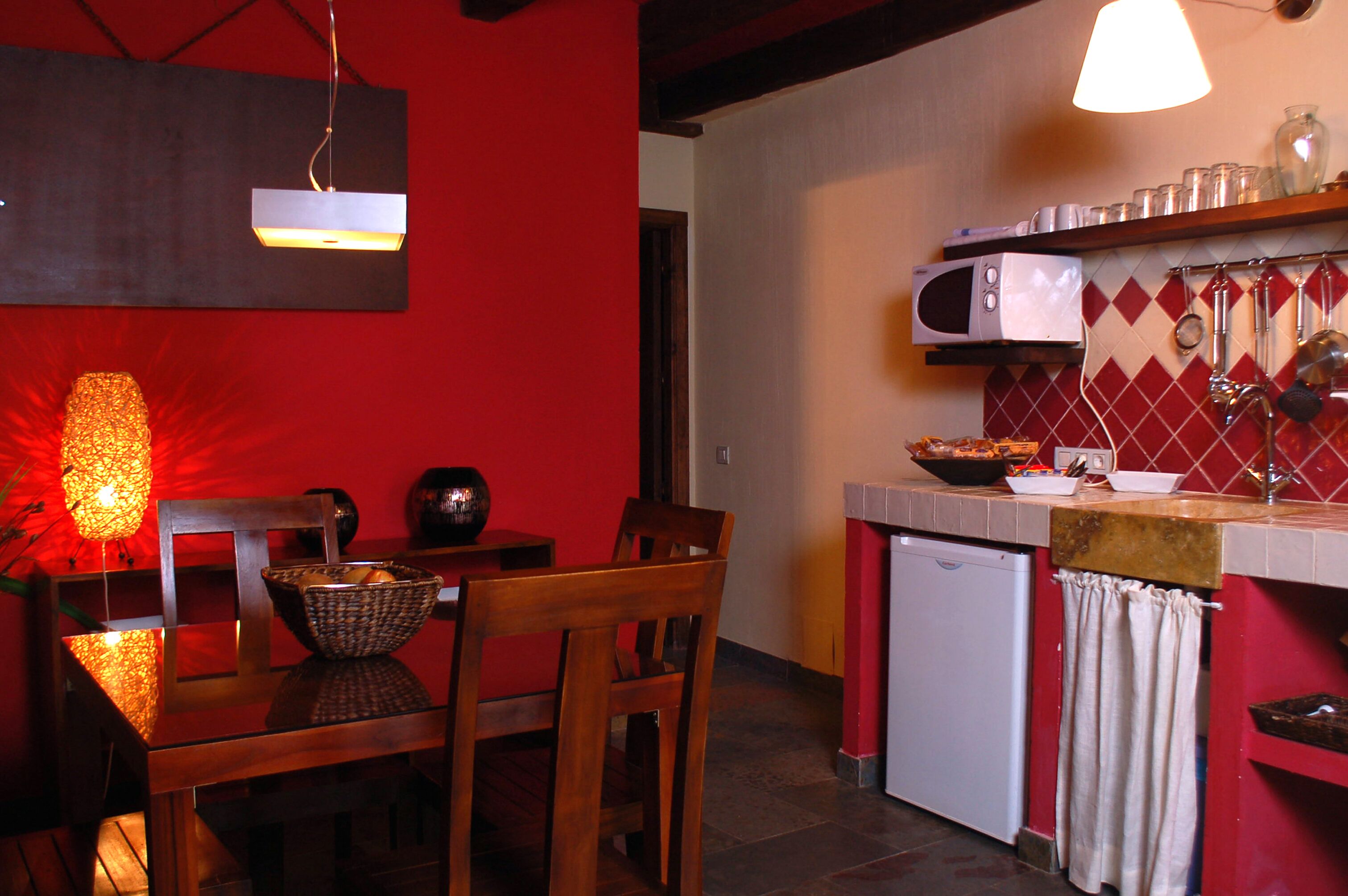 Een kleine keuken met een rode wand, een magnetron, een spoelbak en een eethoek met een tafel en stoelen.