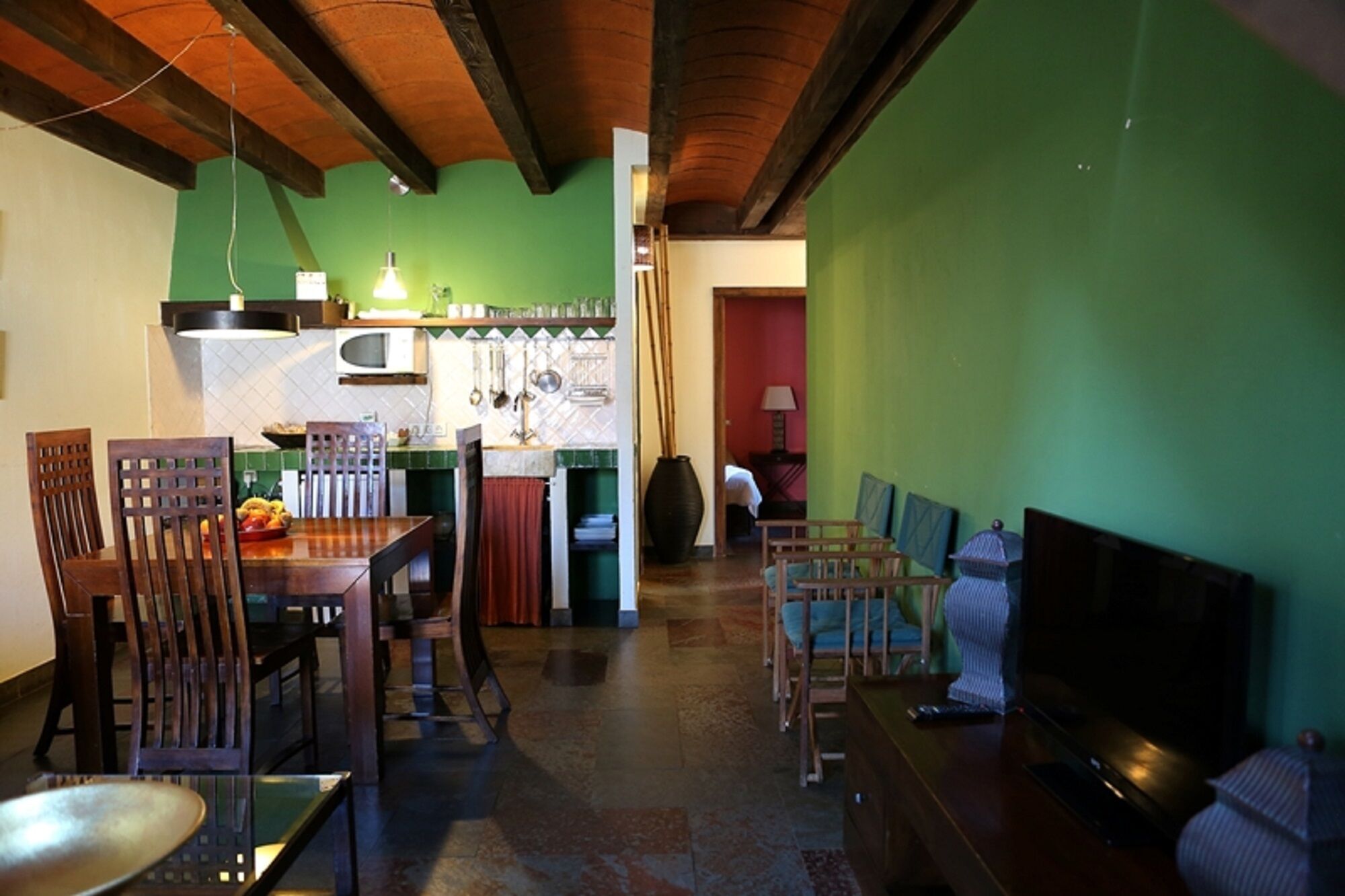 A dining area with wooden chairs and a table, a kitchen with green walls and wooden beams, and a living area with a TV and seating.