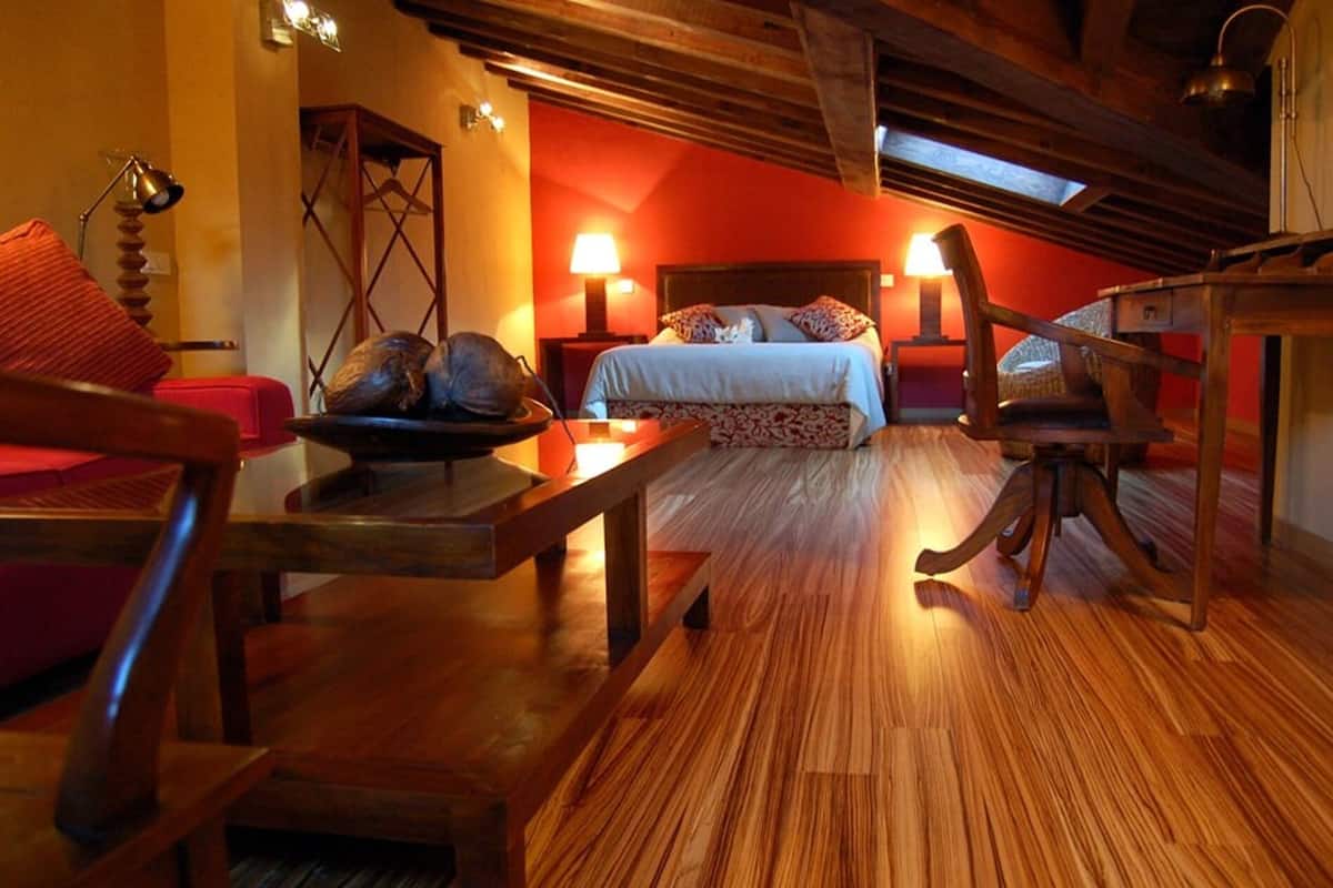A cozy room with a wooden floor, a bed with a patterned bedspread, a red chair, a wooden table with a decorative bowl, and a wooden chair with a curved backrest.