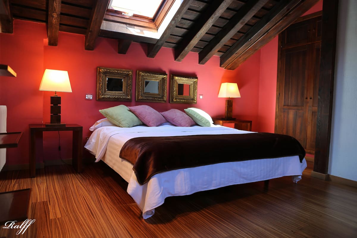 A bedroom with a wooden ceiling, a bed with white bedding and multiple pillows, two bedside lamps, and three framed mirrors on the wall.