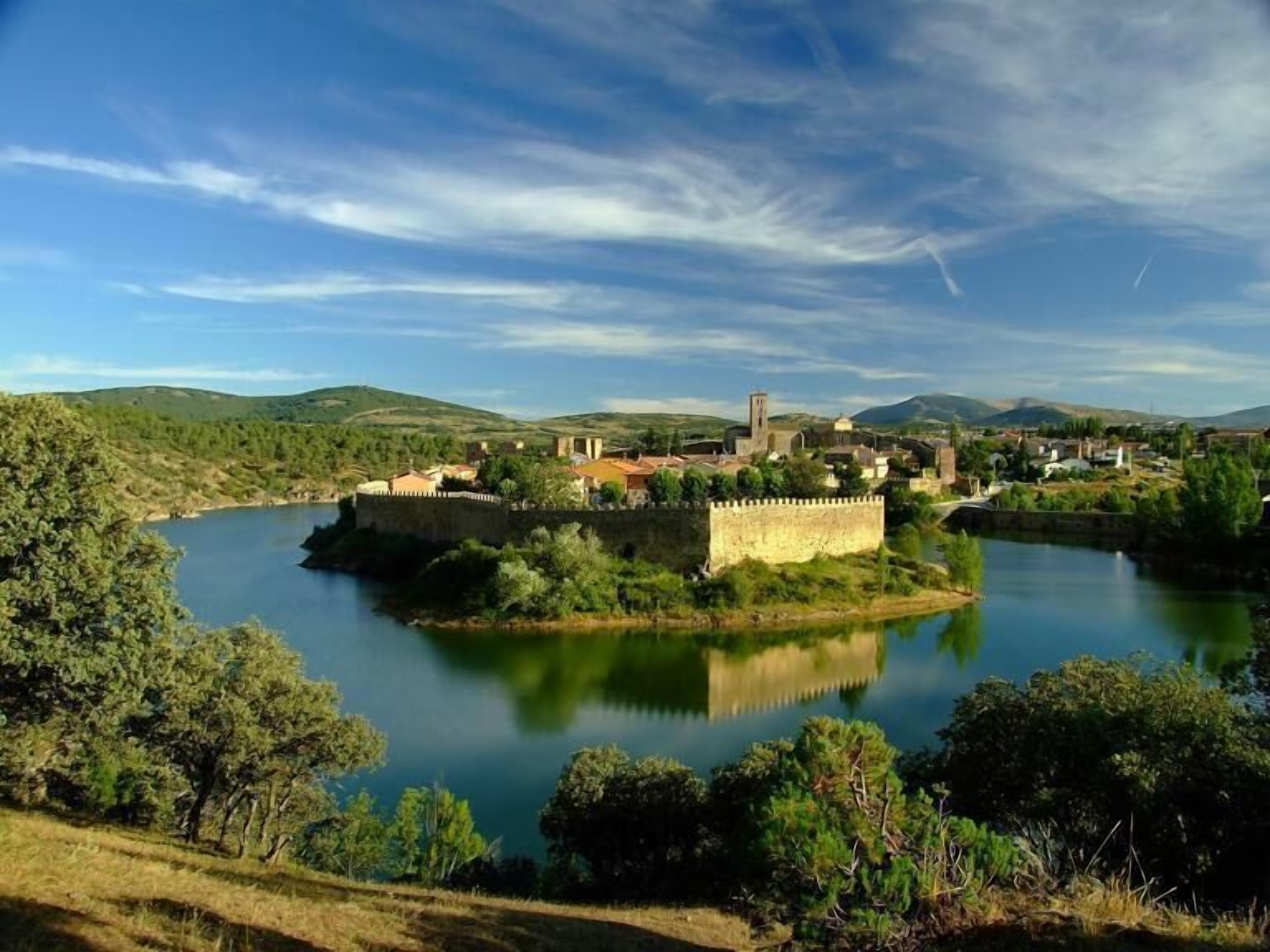 A historic fortification surrounded by a calm river, with lush greenery and distant hills.