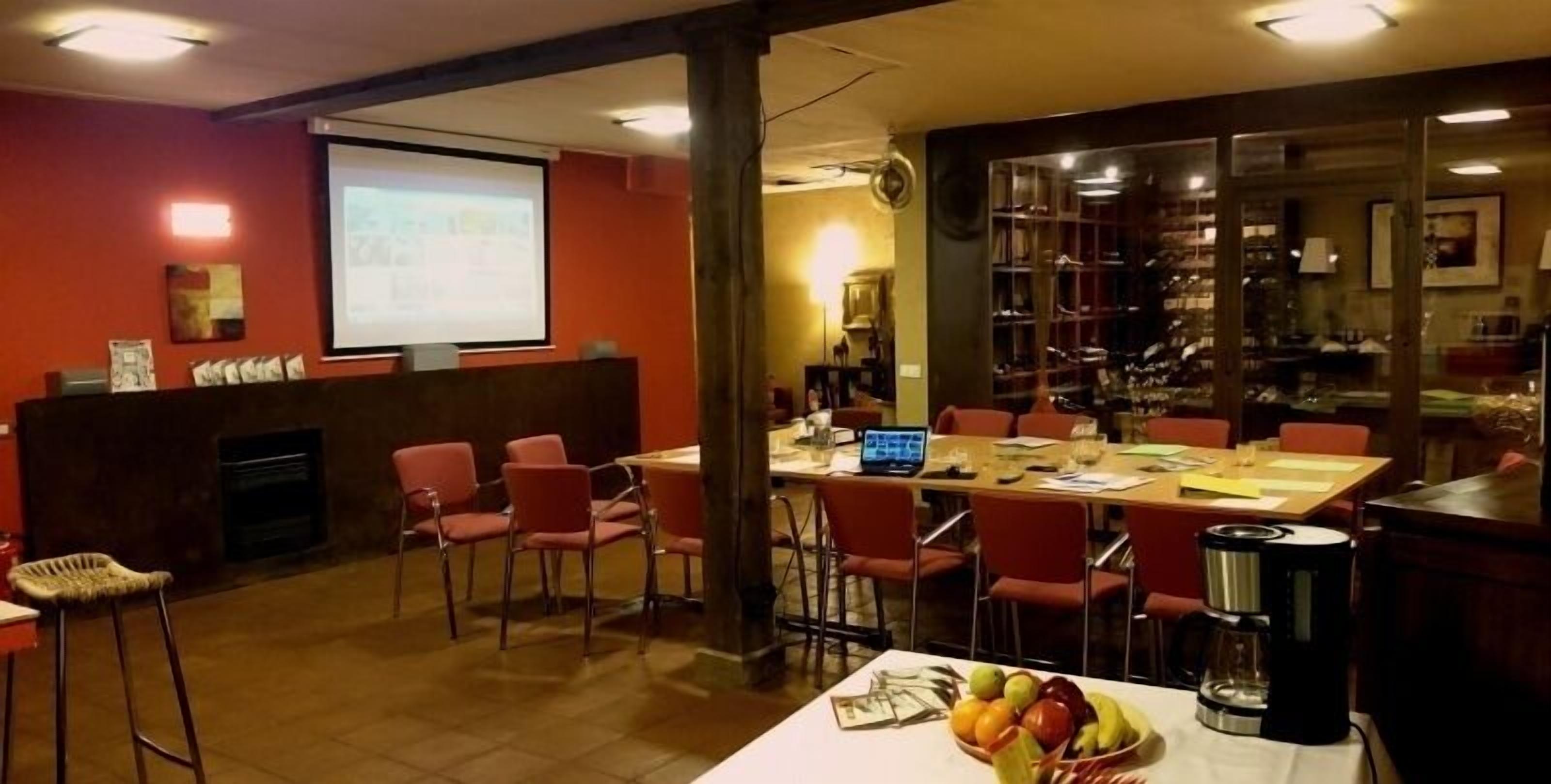 Un salón de conferencias con una larga mesa, sillas rojas, una pantalla de proyector y una máquina de café.