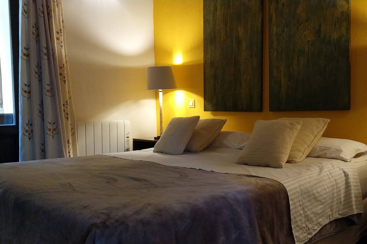 A hotel room with a bed, two bedside lamps, a radiator, and a large painting on the wall.