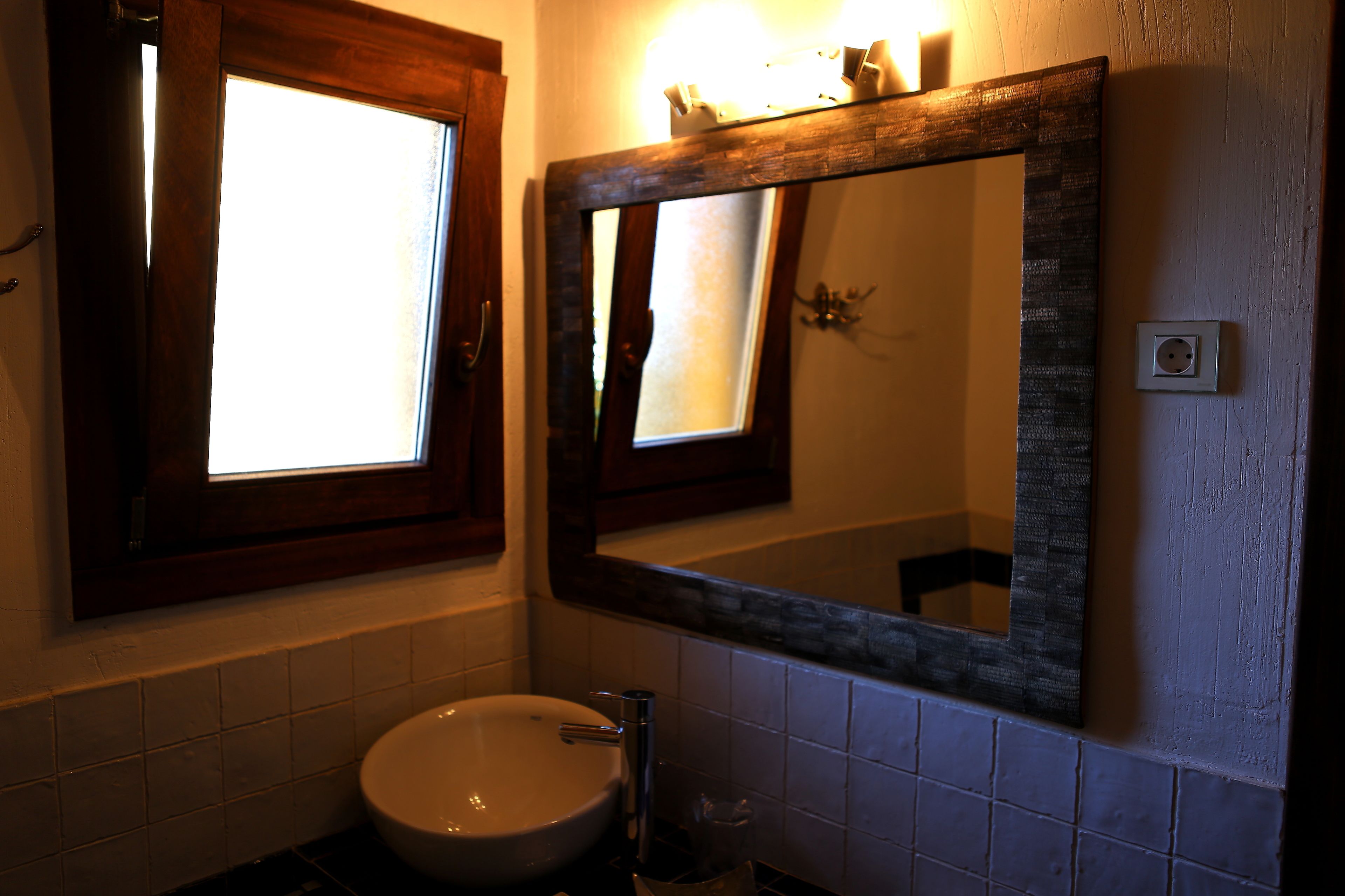 Een badkamer met een witte wastafel, een spiegel met houten lijst en een raam boven de wastafel.