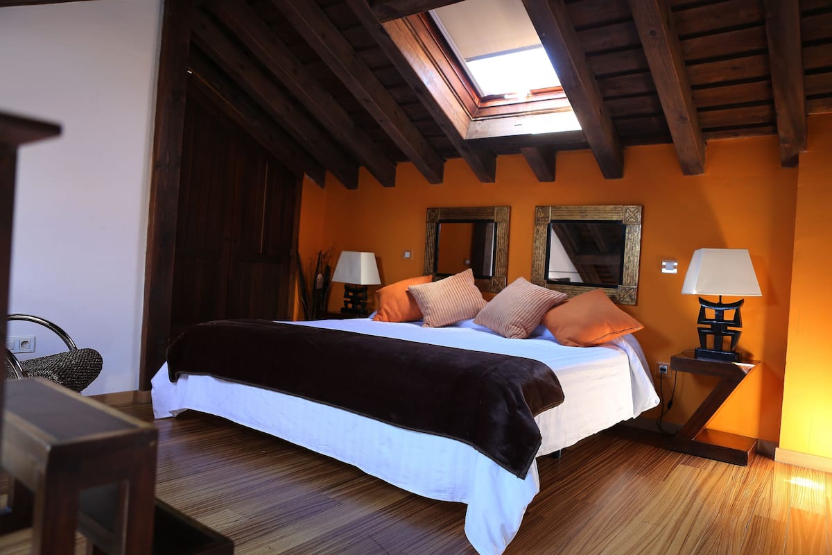 A bedroom with a bed, wooden floor, a skylight, two bedside tables with lamps, and two wall mirrors.