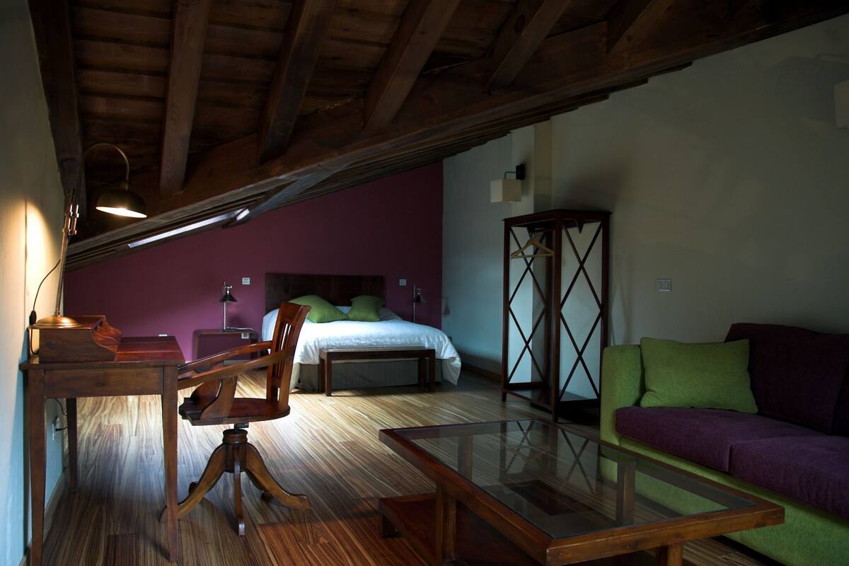 A room with a wooden ceiling, a bed with green pillows, a green sofa, and a glass-top table.