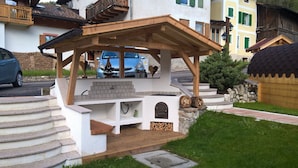 A wooden outdoor kitchen with a stone fireplace, steps leading up to it, and a stone wall in the background. - Residence Le Rose (San Giovanni di Fassa)