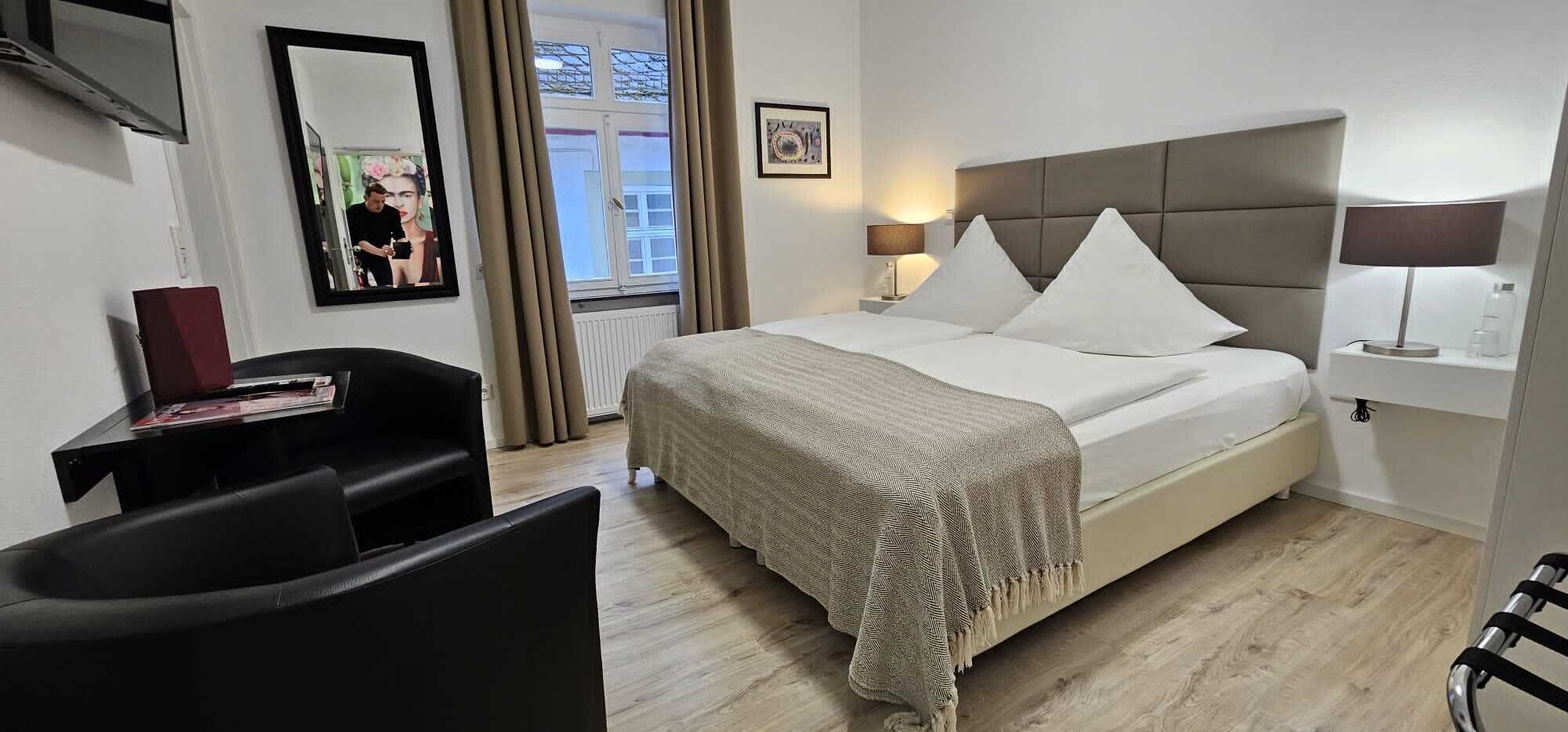 Une chambre d’hôtel comprenant un lit, des lampes de chevet, un bureau avec une chaise, un miroir et une fenêtre avec des rideaux.