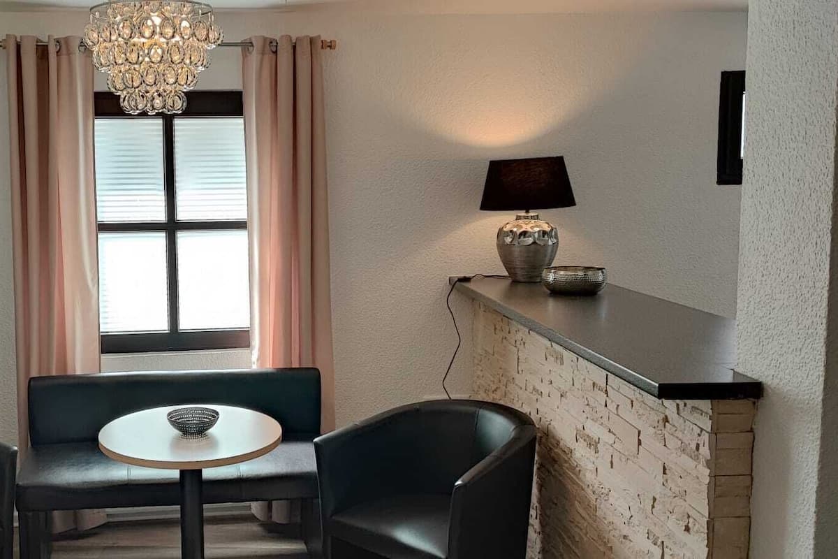 A small room with two black chairs, a round table, a potted plant, a window with curtains, a chandelier, and a lamp on a shelf.