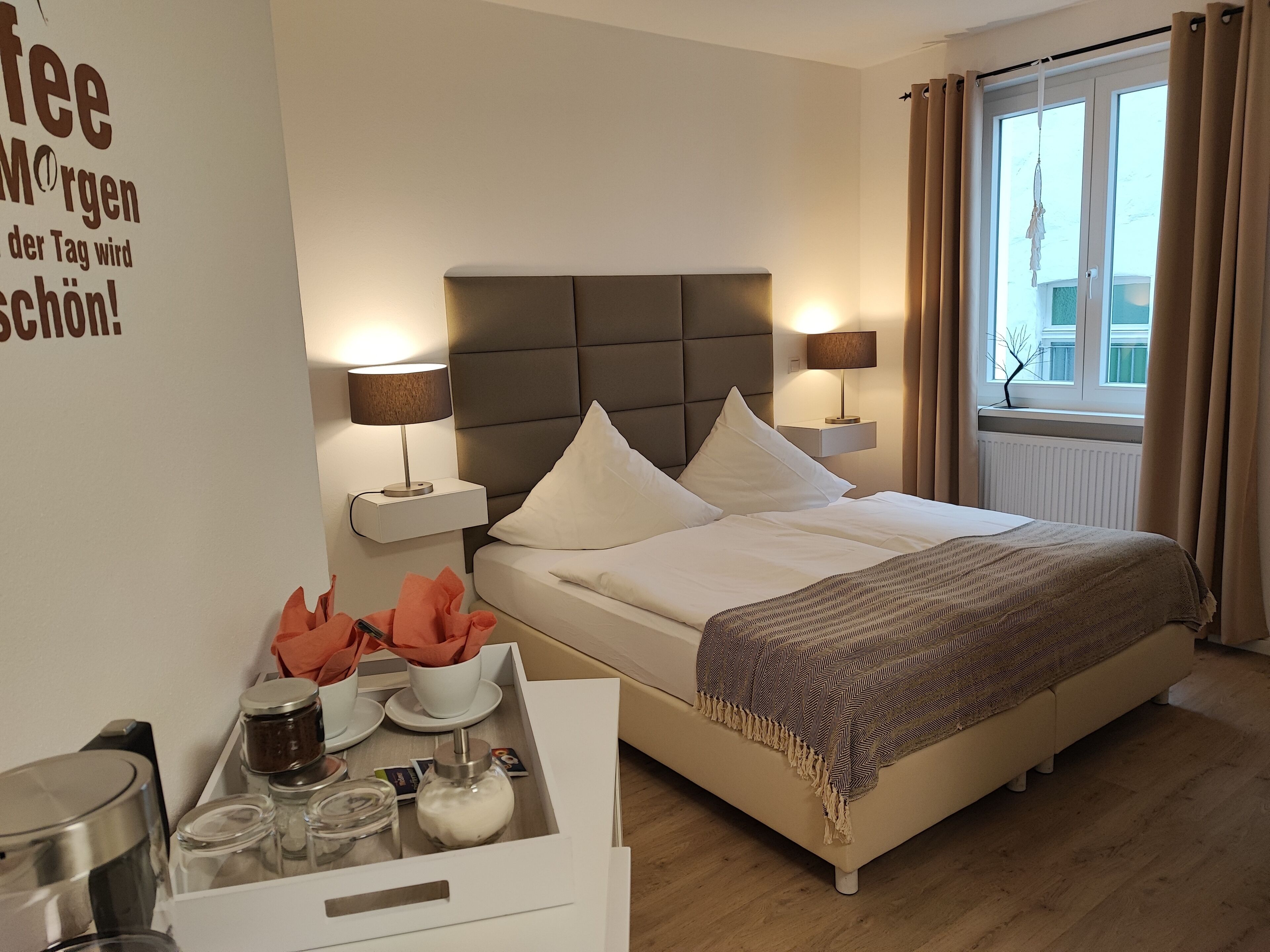 A hotel room with a bed, bedside lamps, a tray with tea and cream, and a window with curtains.
