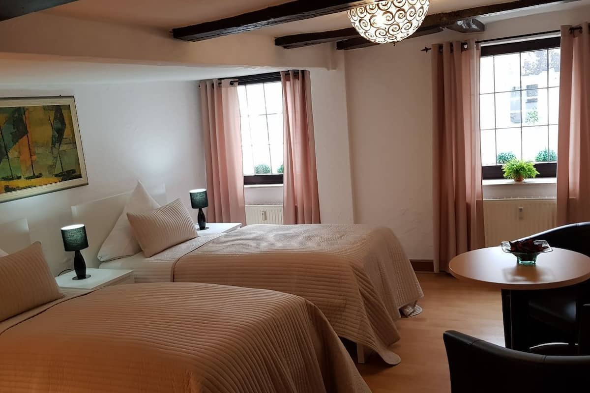 A hotel room with two beds, a small table, a chair, a lamp, and a window with curtains.