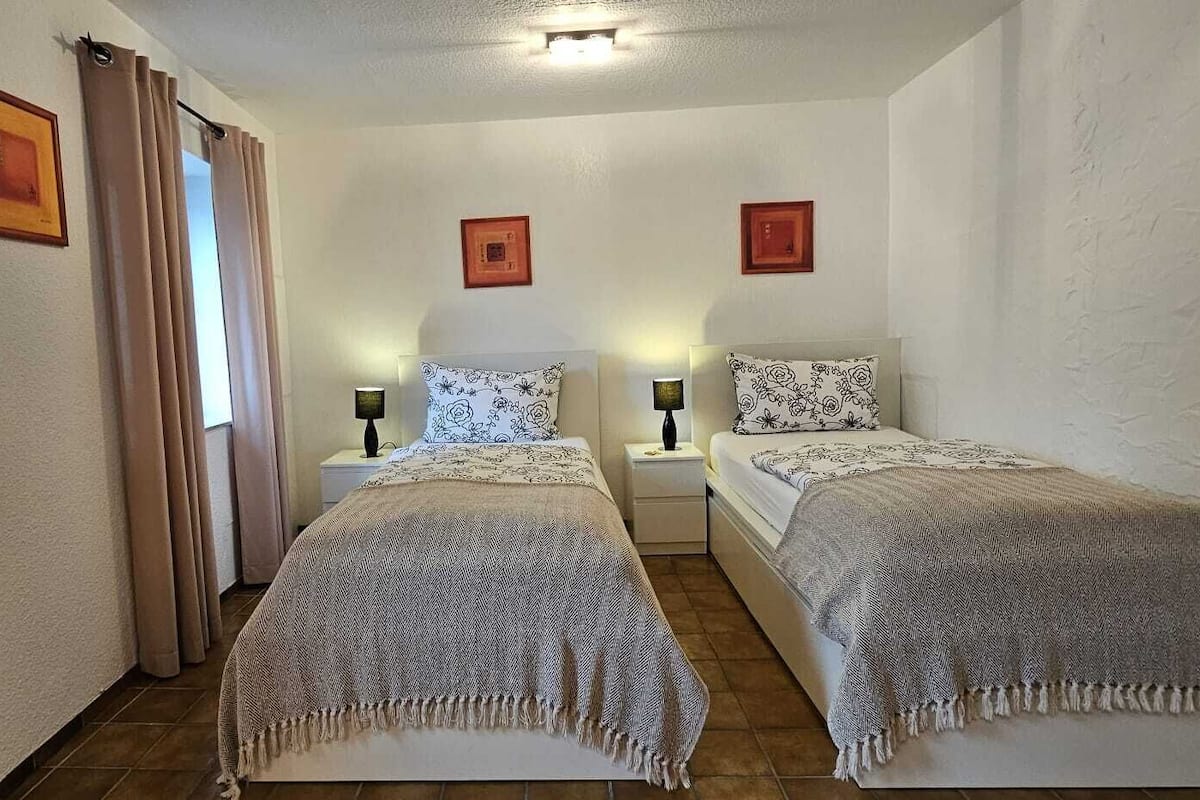 A room with two beds, each with a patterned bedspread and matching pillows. There are two bedside tables with lamps, and the walls are adorned with framed pictures.