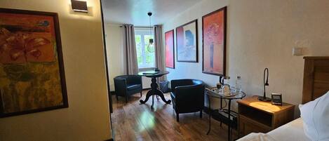 A hotel room with a bed, a desk with a lamp, a chair, a small table with a coffee maker, and artwork on the walls.