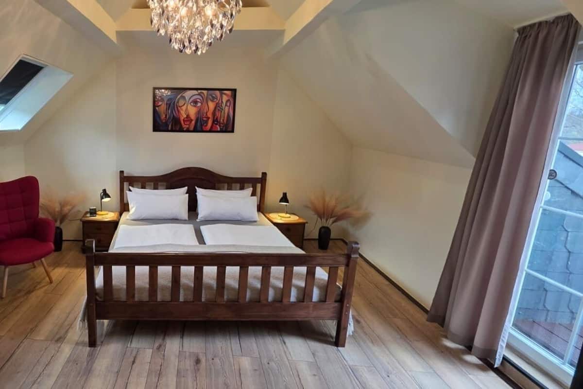 A bedroom with a wooden bed, two red chairs, a small table with a vase, a painting on the wall, and a skylight.
